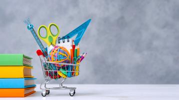 La fotografía tiene un fondo gris. En el lado izquierdo se observan 4 libros cerrados y apilados, a su lado un carrito de compras miniatura y dentro de él hay suministros de manualidades, una tijera, mini regla, lápices y ligas.