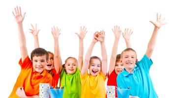 Fotografía en la que aparecen 6 niños y niñas, cada uno con una camisa de color distinto (naranja, naranja oscuro, verde, amarillo, rojo y celeste), todos los niños están sonriendo mientras levantan sus manos.
