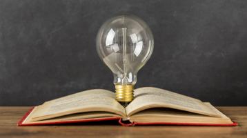 Fondo gris. Sobre una mesa de madera un libro abierto y entre sus páginas un bombillo.