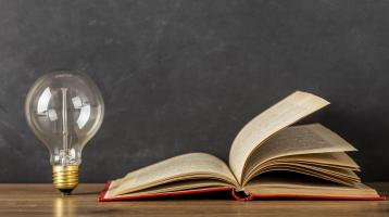 Fondo gris. Sobre una mesa de madera un libro abierto y a la par un bombillo.