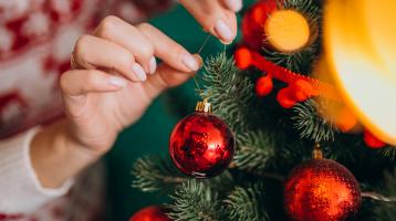 Manos decorando un árbol de navidad con ornamentos circulares rojos. 