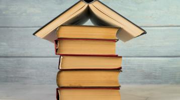 Sobre un fondo de madera celeste, en una mesa una torre de libros y encima un libro abierto formando una casa. 