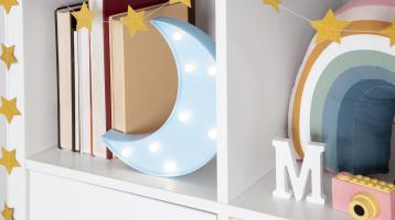 Imagen de un estante con repisas color blanco, decorado con estrellas doradas, en un estante unos libros junto a una figura de luna con luz, en el otro estante se observa una letra M, una cámara de juguete y un almohadón en forma de arcoiris.
