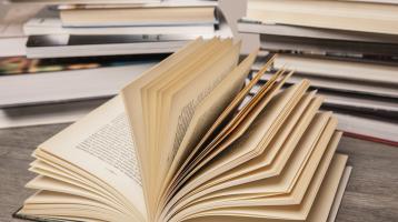 Sobre una mesa de madera, un libro abierto al frente, y atrás una pila de libros. 