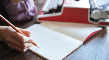 Sobre una mesa de madera, una persona escribiendo en un libro y a la par una máquina de escribir.
