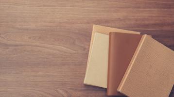 Sobre una mesa de madera, en la esquina inferior derecha 4 libros apilados de color café.