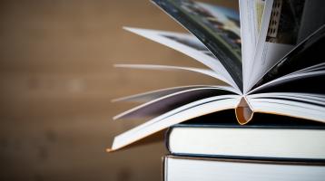 Fondo de madera. A la izquierda una torre de libros y en encima un libro abierto.