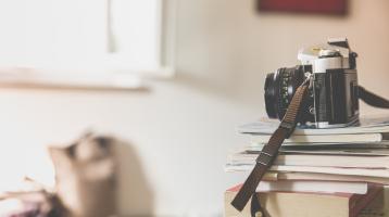 Al fondo una ventana y unos objetos desenfocados en primer plano una cámara fotográfica sobre unos libros