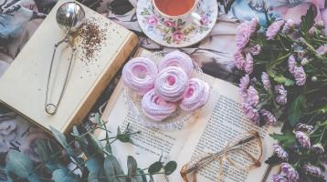 Varios objetos vistos desde arriba, 2 libros uno abierto y otros cerrado, unas flores, una taza conté y unos lentes.